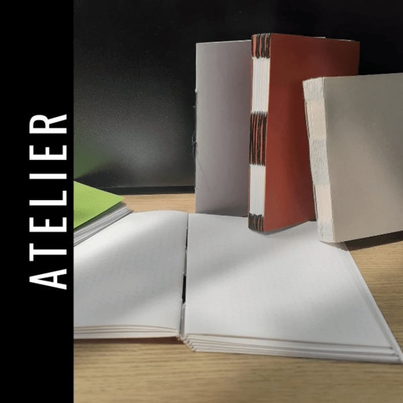 In the foreground, an open white notebook lies on a wooden surface, while in the background, three bound books stand against a dark backdrop.
