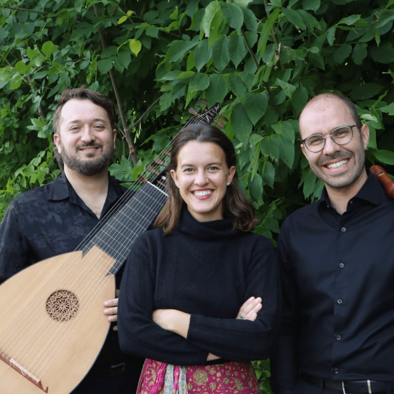 Three smiling musicians, two with instruments, stand in front of lush green foliage.