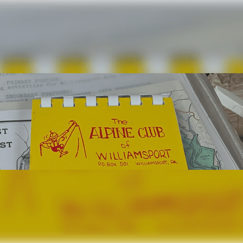 A yellow card with "The ALPINE CLUB of WILLIAMSPORT" printed in red is in the foreground, with papers and a map visible in the background.