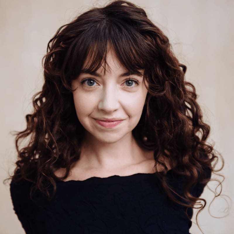 A young woman with curly brown hair and blue eyes smiles gently in the foreground against a softly lit, neutral background.