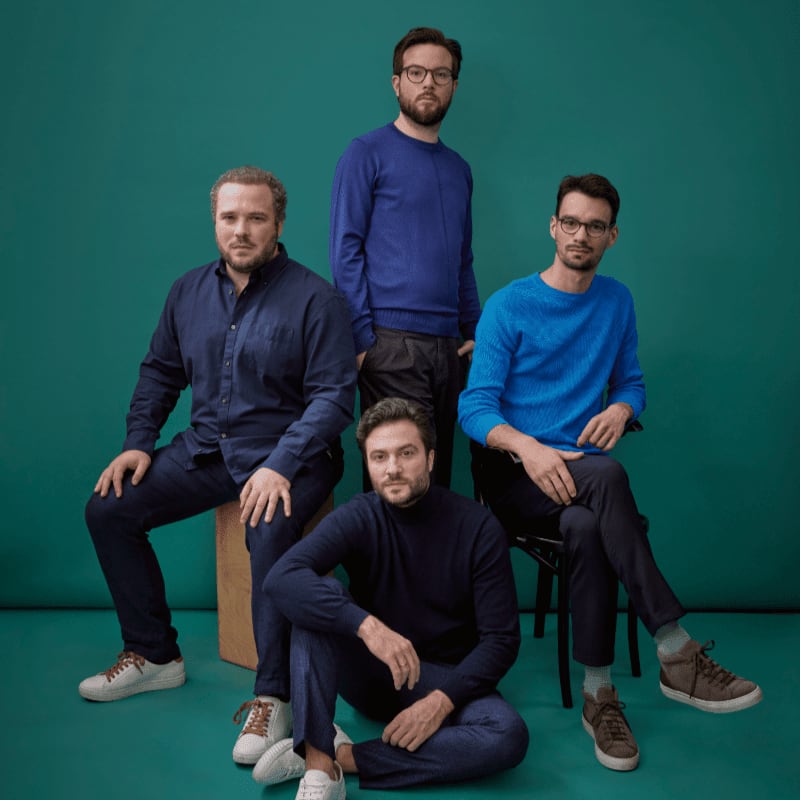 Four men in blue sweaters and shirts pose against a teal background, with one man sitting on the floor in the foreground.
