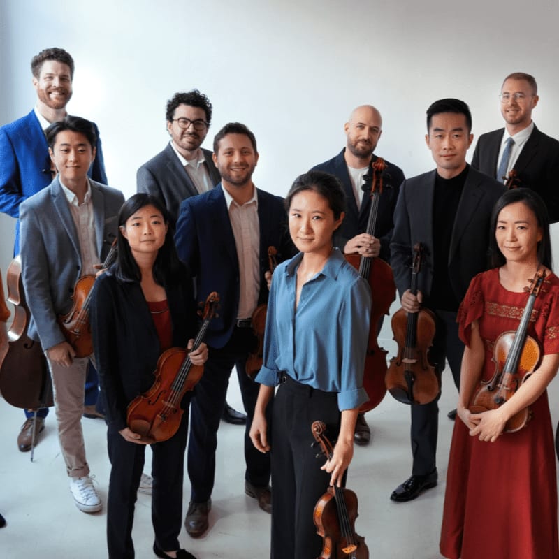 A group of musicians in formal attire stand with their instruments in front of a plain white background.