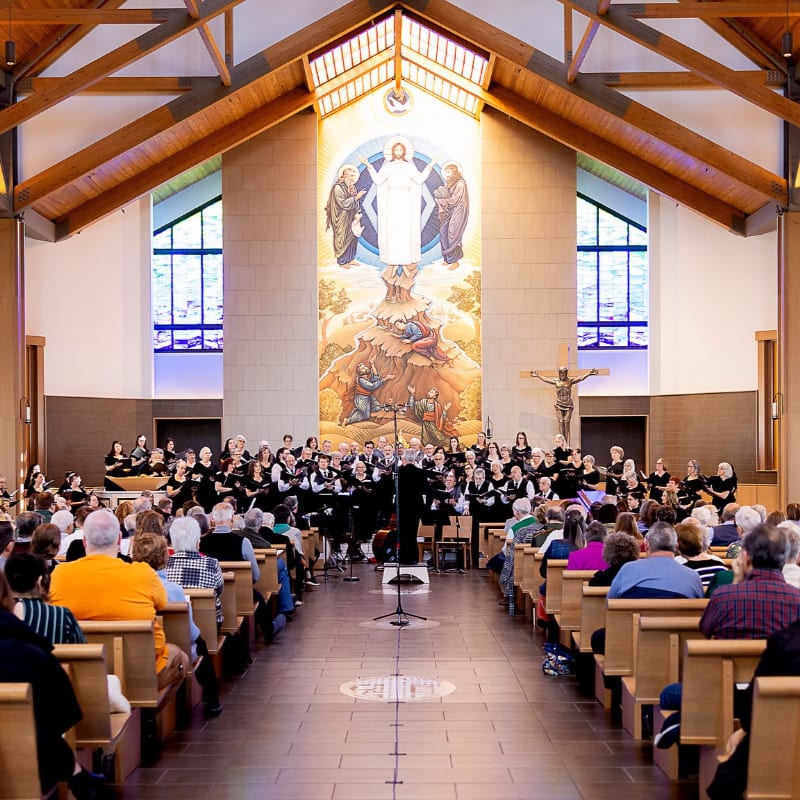 A choir performs in a church with a large mural of Jesus Christ behind them and an audience seated in pews in the foreground.