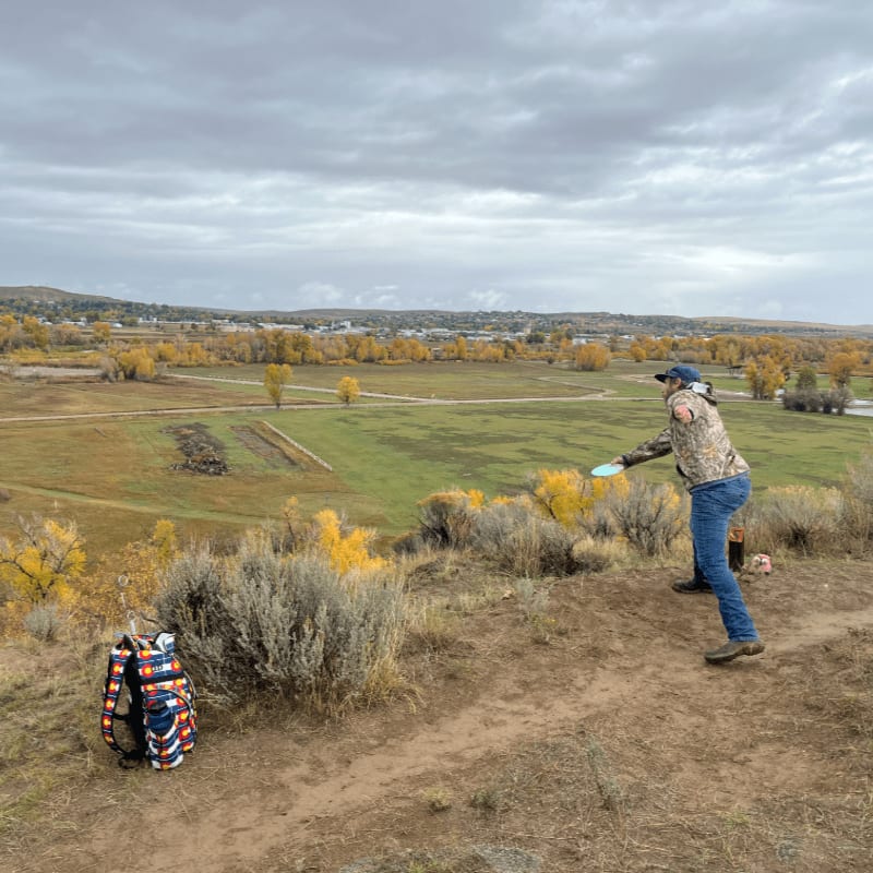 A person throws a frisbee on a grassy hill in the foreground, with a town and autumn trees in the background under a cloudy sky.