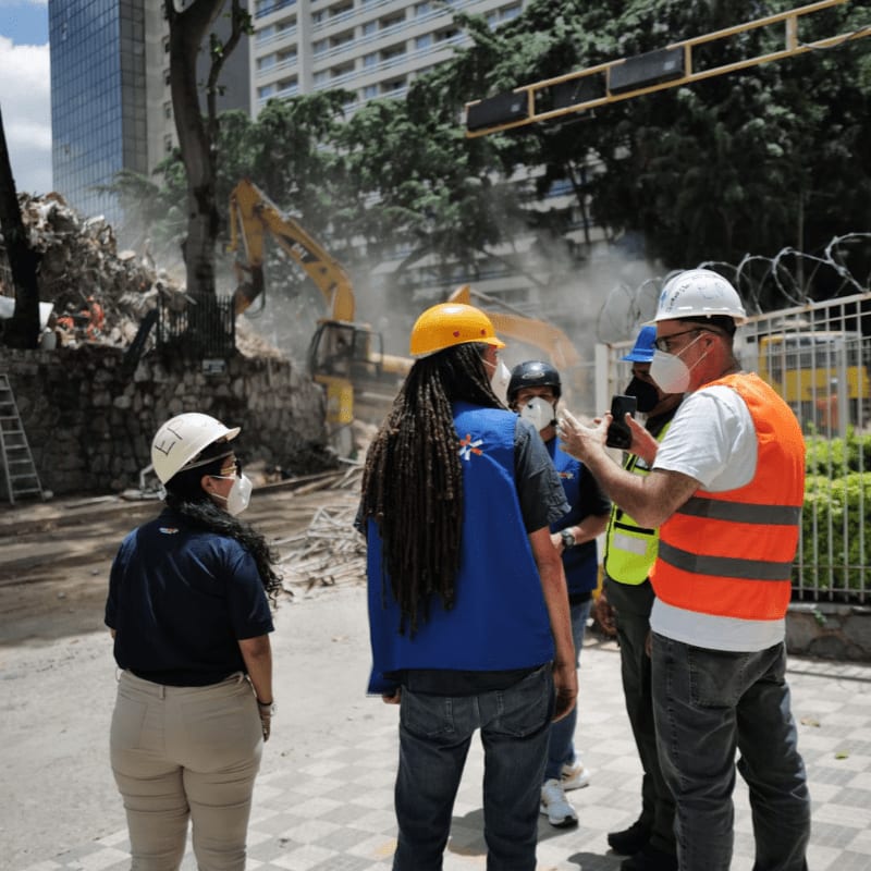 Four people in hard hats and safety vests stand in the foreground, observing a construction site with excavators and debris in the background.
