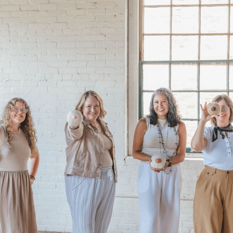 Four women stand in front of a white brick wall and a large window, holding decorative breast-shaped objects.