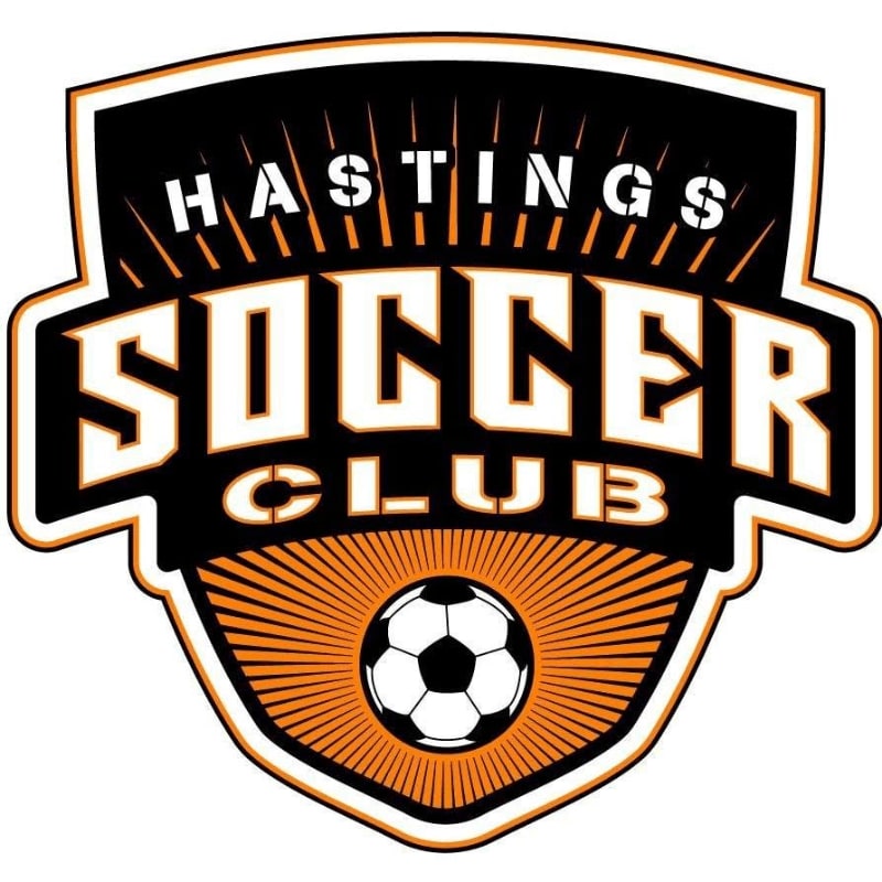 A soccer ball is centered in the foreground of a shield-shaped logo with "HASTINGS SOCCER CLUB" written in bold letters against a black and orange background.