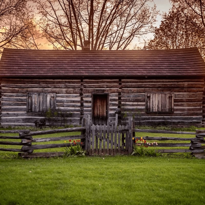 A rustic log cabin with a wooden fence and green grass in the foreground is set against a backdrop of bare trees and a warm, hazy sky.