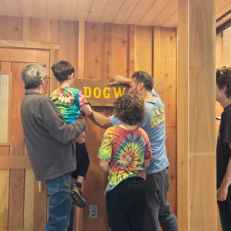 Several people, including a child in a tie-dye shirt, are hanging a "DOGWOOD" sign on a wooden wall in a room with wooden paneling and a door.