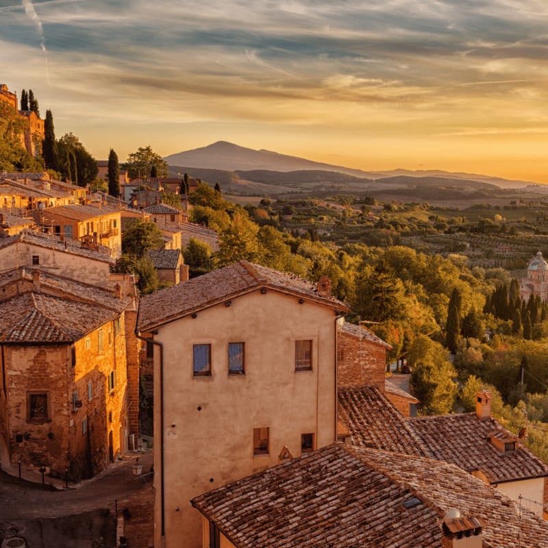 Tuscan rooftops and rolling green hills are bathed in the warm glow of a sunset, with a distant mountain peak silhouetted against the sky.