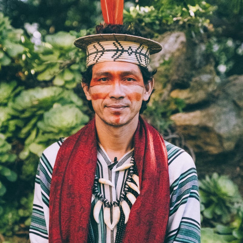 A man with face paint and traditional attire stands in front of lush green foliage.