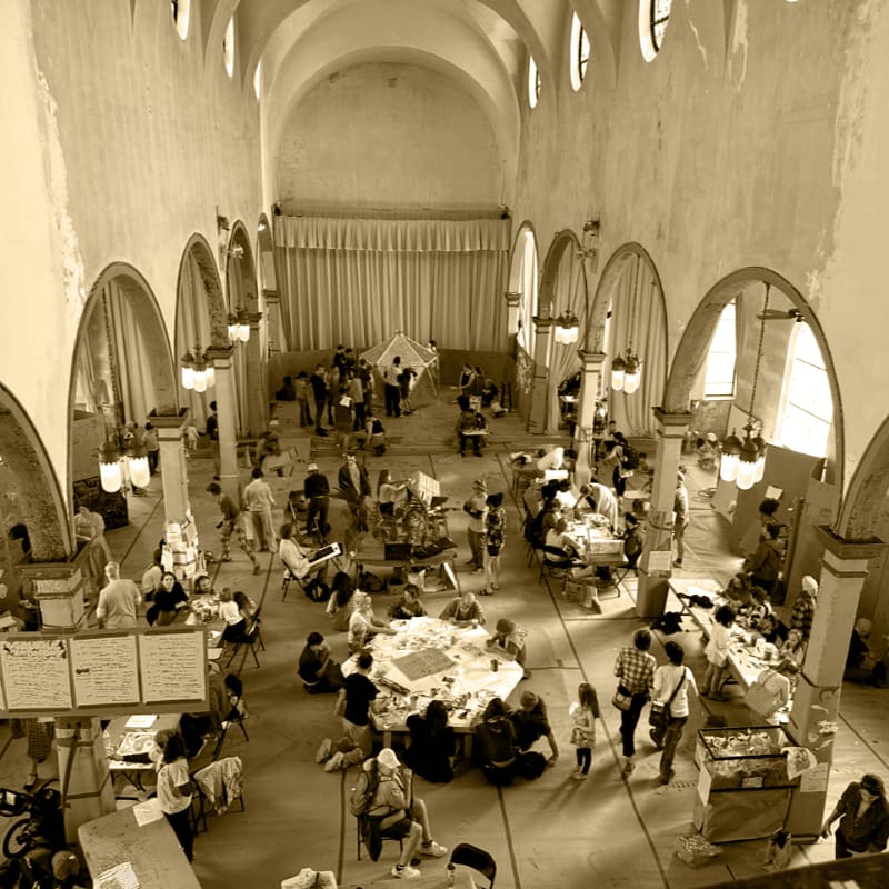 People gather around tables in a large, arched hall with a stage and curtains in the background.