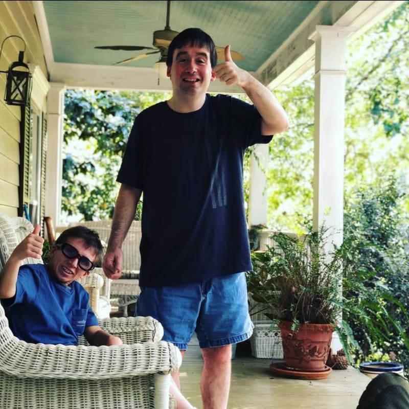 Two men give thumbs up on a porch with lush greenery in the background.