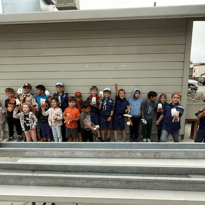 A group of children in blue uniforms stand in front of a beige wall, holding white objects, with a street visible in the background.