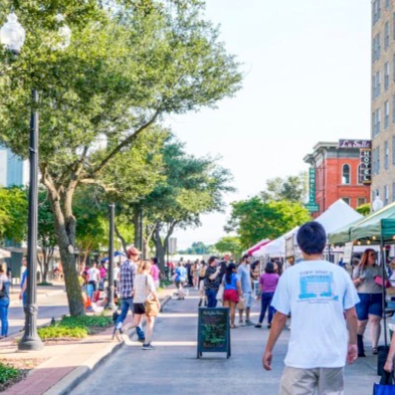 People stroll along a sunny street lined with trees and market stalls, with a tall building visible in the background.