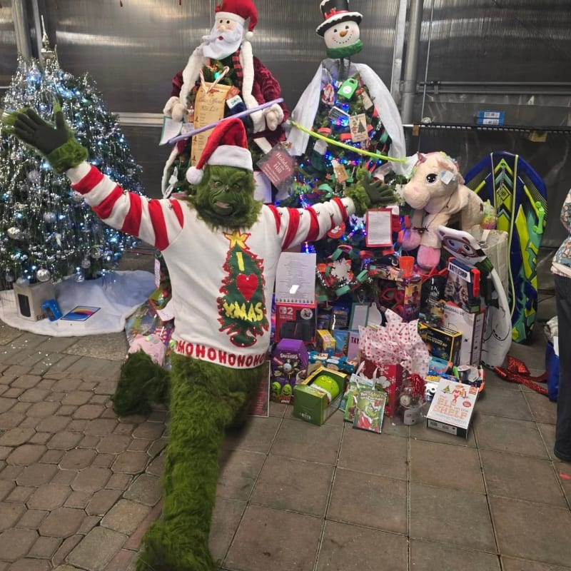 A Grinch character in a festive sweater poses in front of a pile of wrapped gifts, a decorated Christmas tree, and two large figures of Santa Claus and a snowman.