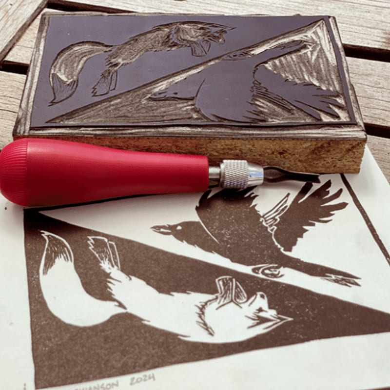 A linocut block with a carved fox and bird design rests above a freshly printed impression of the same design, with a carving tool in the foreground.
