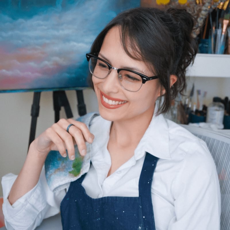 A smiling woman in glasses and an apron holds a paint-stained cloth in the foreground, with an easel and art supplies visible in the background.