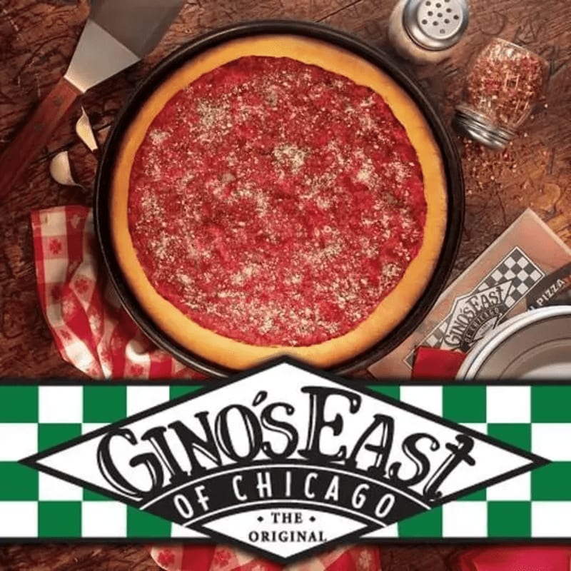 A deep-dish pizza with a thick crust and red sauce sits on a wooden table with a spatula and garlic in the foreground, while a Gino's East of Chicago logo is prominently displayed at the bottom.
