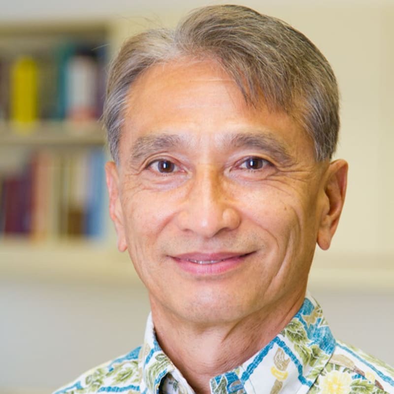 A smiling East Asian man with graying hair wears a floral shirt in the foreground, while a blurred bookshelf fills the background.