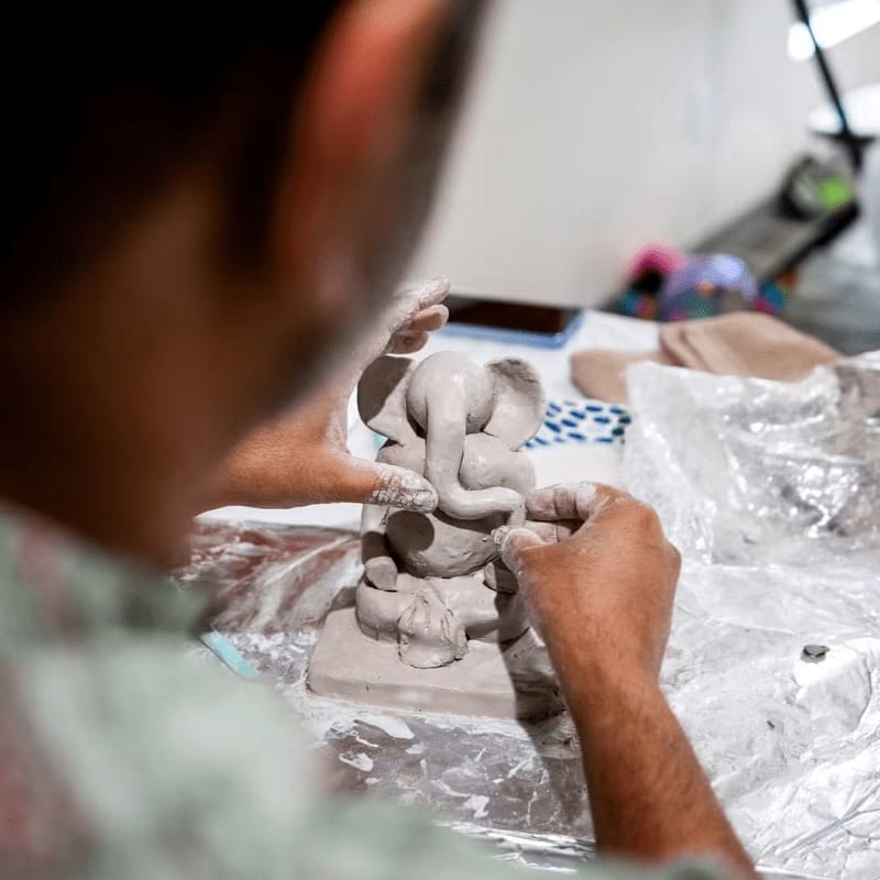 Hands sculpt a clay Ganesha figure in the foreground, with a blurred background of crafting materials and a phone.