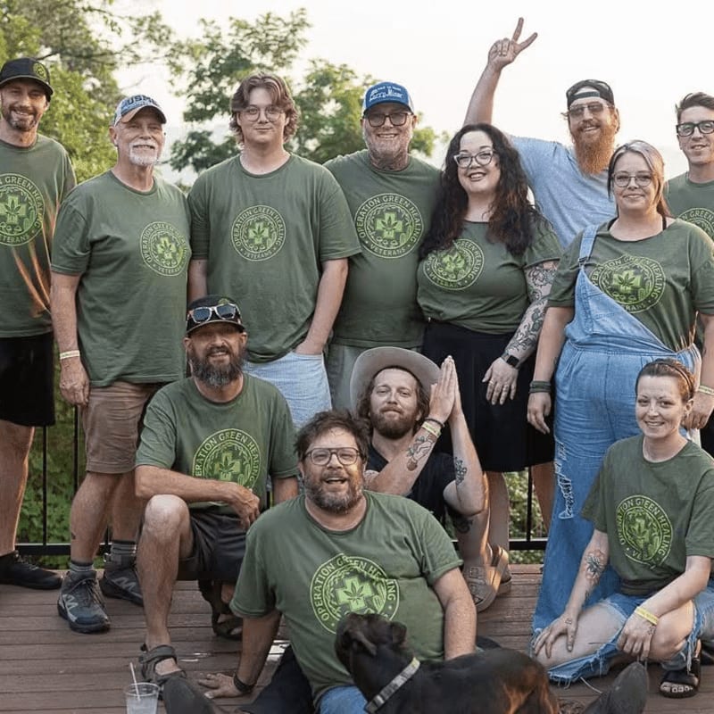 A group of people in matching green shirts pose for a photo on a wooden deck with a dog in the foreground and trees in the background.