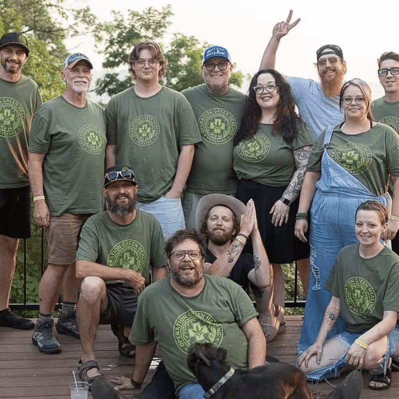 A group of people in matching green shirts pose for a photo on a wooden deck with a dog in the foreground and trees in the background.