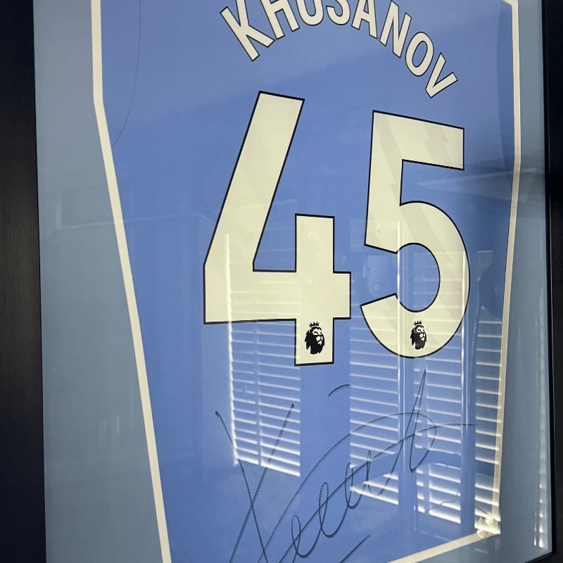 A framed blue jersey with the name "KHUSANOV" and the number "45" is displayed against a background of blinds reflecting light.
