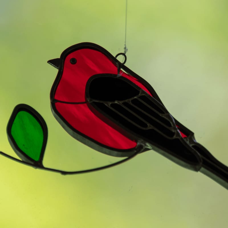 A stained glass bird with red and black plumage perches on a branch with a green leaf, set against a soft, blurred green background.