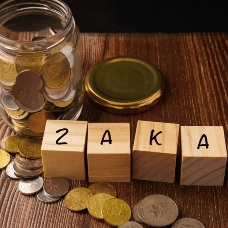 A glass jar filled with coins sits beside four wooden blocks spelling "ZAKA" on a wooden surface, with scattered coins in the foreground and a golden lid in the background.