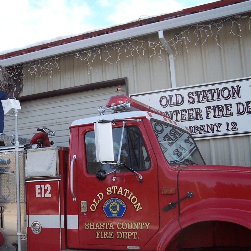 A red fire truck with "Old Station Shasta County Fire Dept." emblazoned on its side is parked in front of a building with "Old Station Volunteer Fire Dept. Company 12" signage and festive lights.