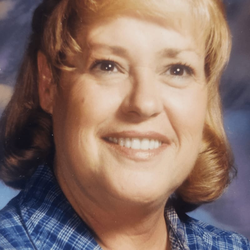 A smiling woman with blonde hair and a blue plaid shirt is in the foreground, with a blurred blue and white background.