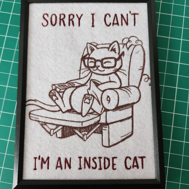 A framed embroidery of a cat wearing glasses and holding a remote control while relaxing in a recliner, with the text "SORRY I CAN'T I'M AN INSIDE CAT" above and below, is set against a green grid background.
