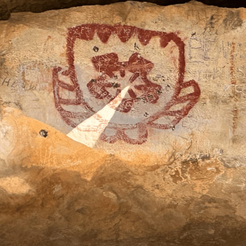 A red, shield-like petroglyph with intricate designs is painted on a rough, textured rock face, with faint scratches and markings visible in the background.