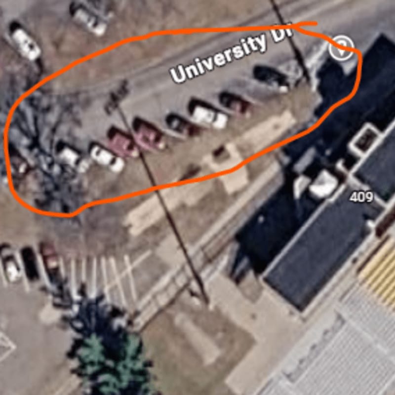 Cars are parked along University Drive in the foreground, with a building and trees visible in the background.