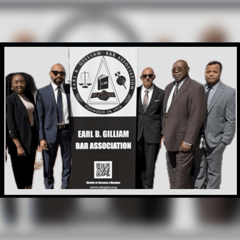 Five formally dressed individuals stand in front of a large sign for the Earl B. Gilliam Bar Association, with a bright sky visible in the background.