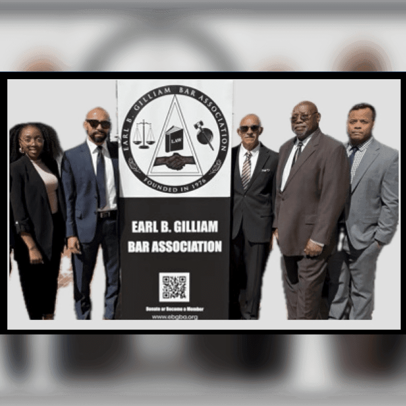 Five formally dressed individuals stand in front of a large sign for the Earl B. Gilliam Bar Association, with a bright sky visible in the background.