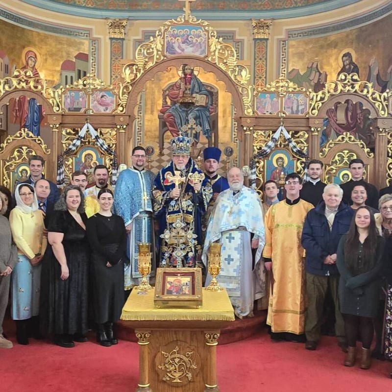 A group of people in religious attire and civilian clothes stand in front of an ornate altar with icons and religious paintings in the background.