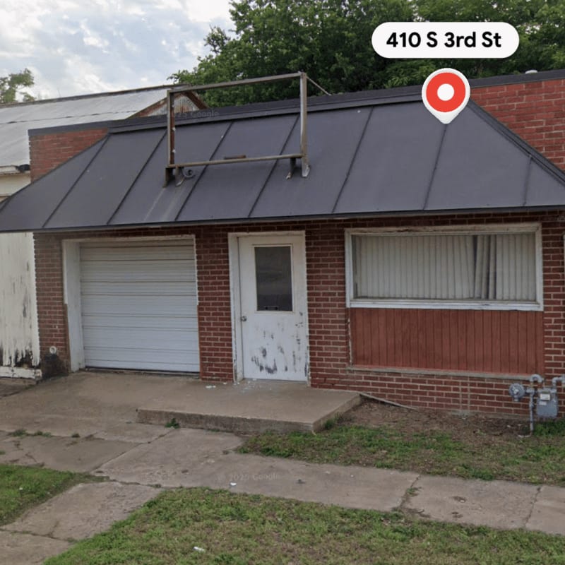 A weathered brick building with a roll-up garage door and a single door stands on a cracked concrete lot, backed by lush green trees.