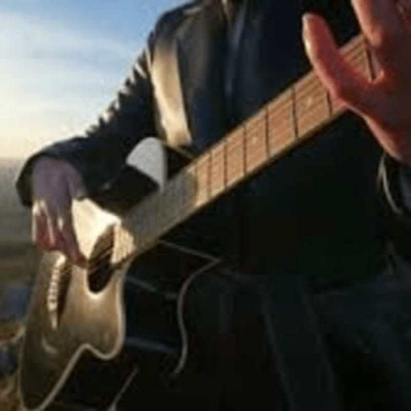A person in a dark jacket plays an acoustic guitar outdoors with a hazy sky in the background.