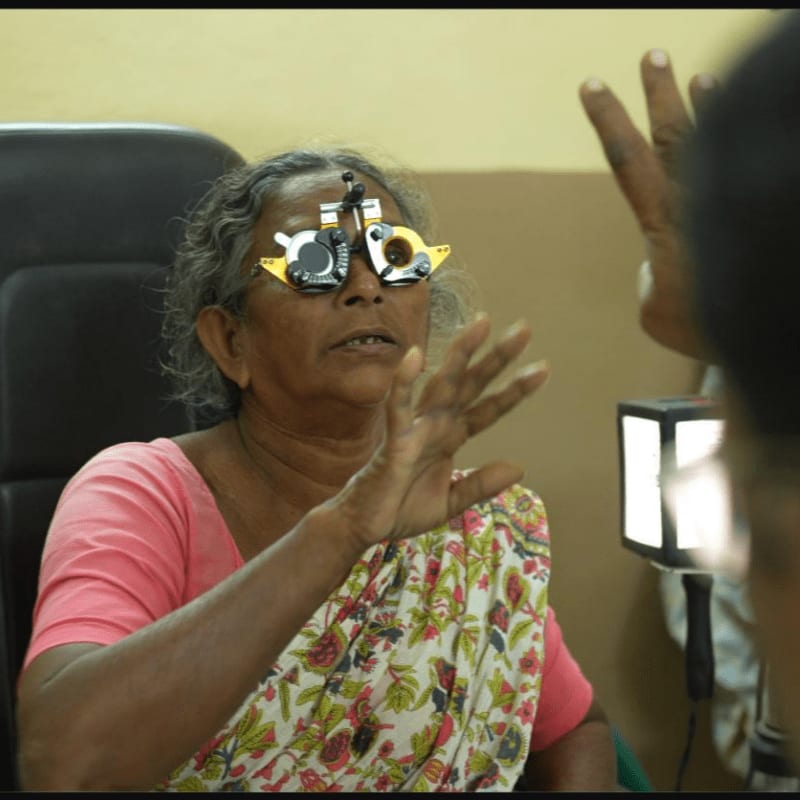 An elderly woman with a trial frame on her eyes reaches out her hands in a dimly lit room with a bright light source in the background.
