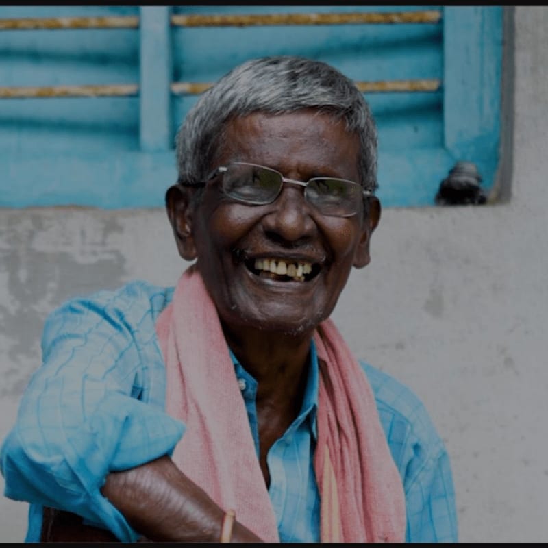 A smiling elderly man with glasses and a pink scarf is seated in front of a blue-framed window.