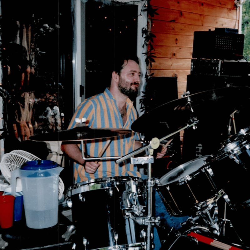A man plays a drum set in the foreground, with a wooden building and speakers in the background.
