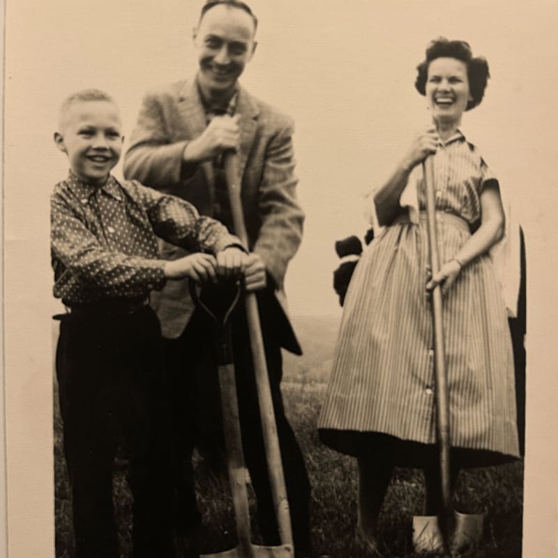 A smiling family of three, a boy and his parents, stand with shovels in a grassy field under a pale sky.