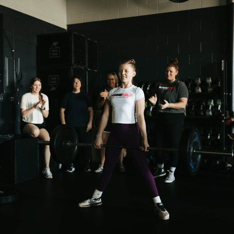 A young woman in the foreground prepares to lift a barbell while a group of women in the background clap and cheer in a gym.