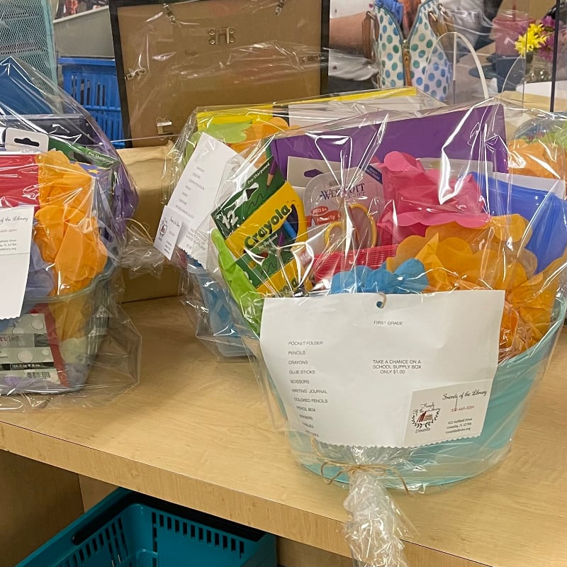 Two gift baskets filled with school supplies and colorful decorations sit on a wooden shelf, with more items and a framed picture visible in the background.