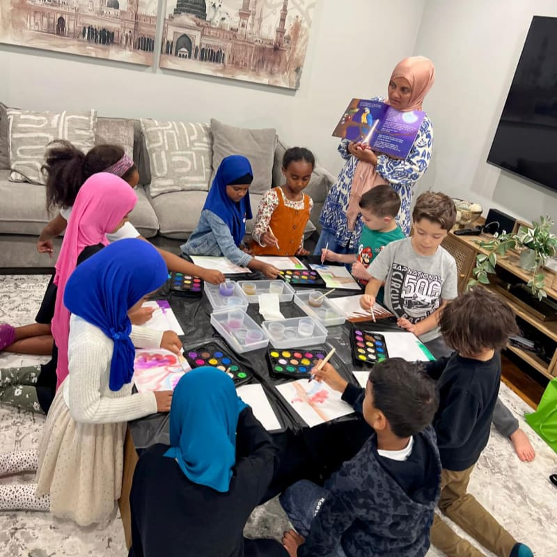 Children are gathered around a table painting and listening to a woman read a book, with a large painting of a mosque on the wall behind them.