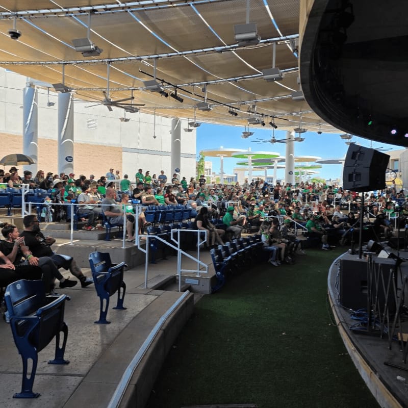 Empty blue seats in the foreground face a large crowd gathered on green turf in front of a stage under a covered outdoor amphitheater.