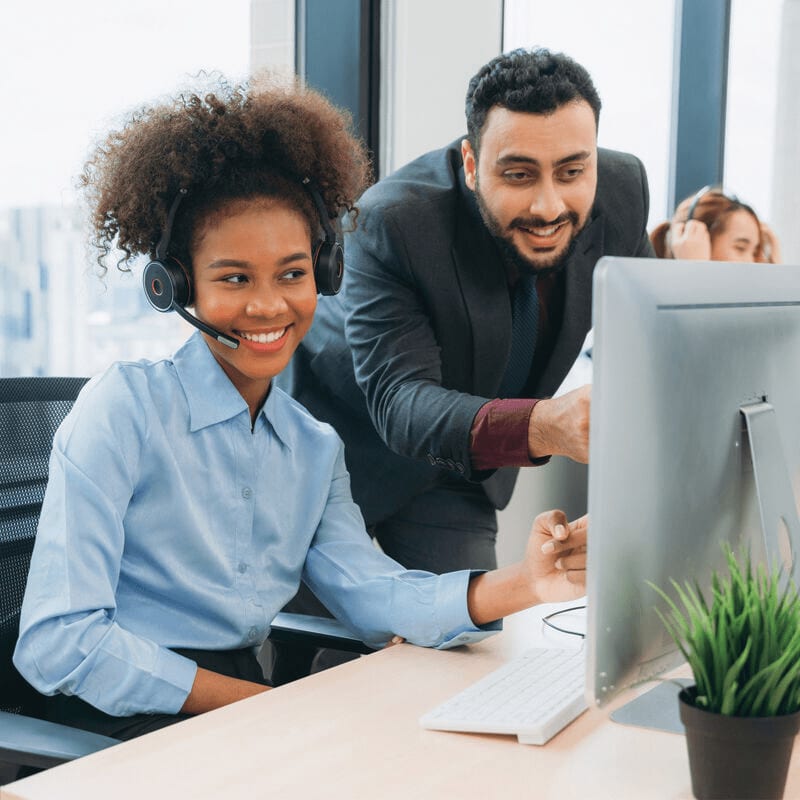 A young woman with a headset smiles while a man points at a computer screen, with other colleagues working in the blurred background.