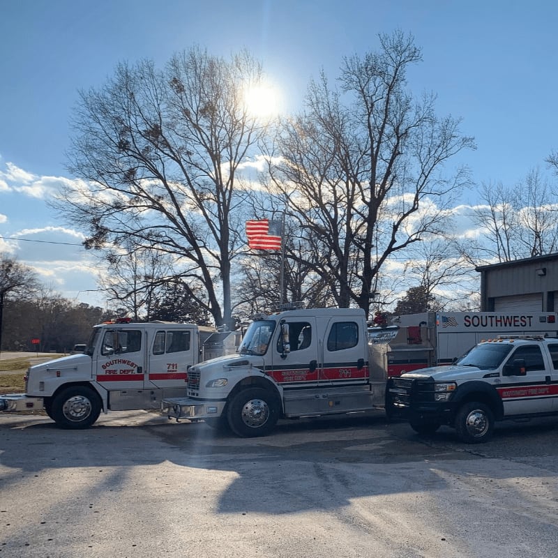 Three white fire trucks are parked in front of a building with an American flag flying above bare trees in the background.